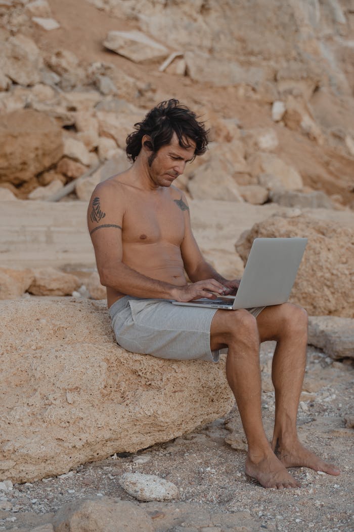 Shirtless man using laptop on a rocky beach, embodying digital nomad lifestyle.